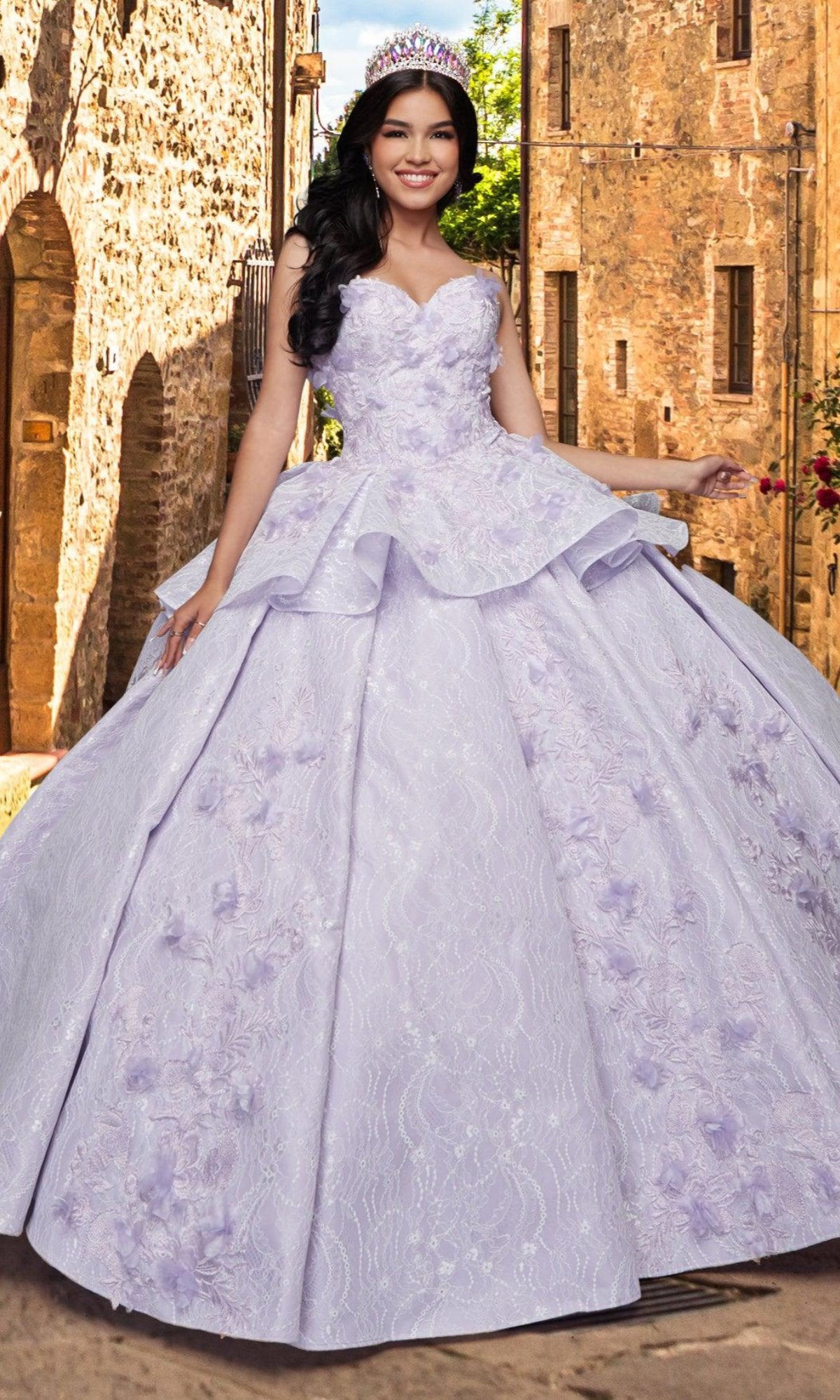 A young woman wearing the Quinceañera Sample Dress QF115 and a tiara smiles on a cobblestone street, stone buildings in the background.